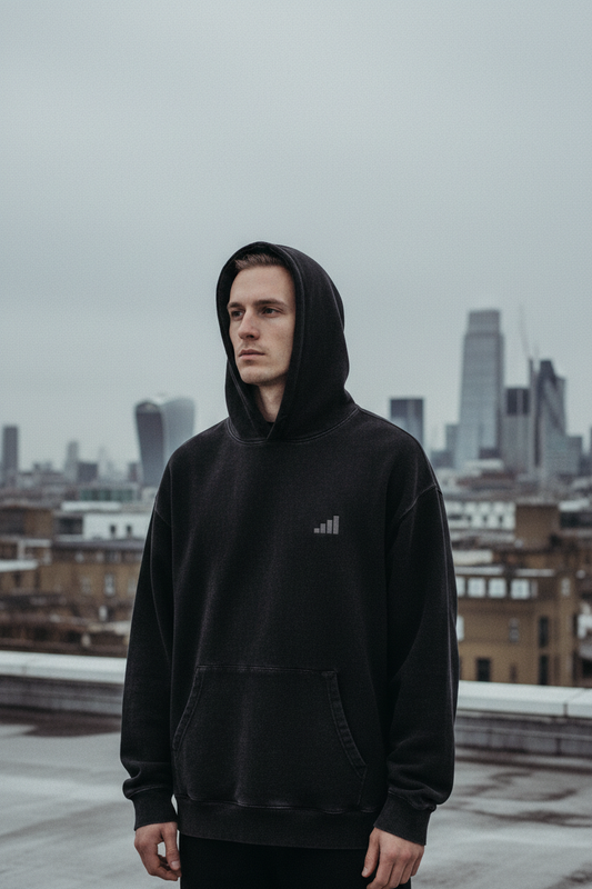 light-skinned male figure wearing washed black aegis. heavyweight hoodie on Bethnal Green rooftop, London skyline behind, overcast grey sky, cinematic editorial