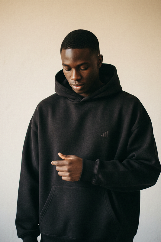 dark-skinned male figure wearing washed black aegis. heavyweight hoodie feeling the fabric weight between fingers, four-bar tonal embroidery on left chest, warm studio background